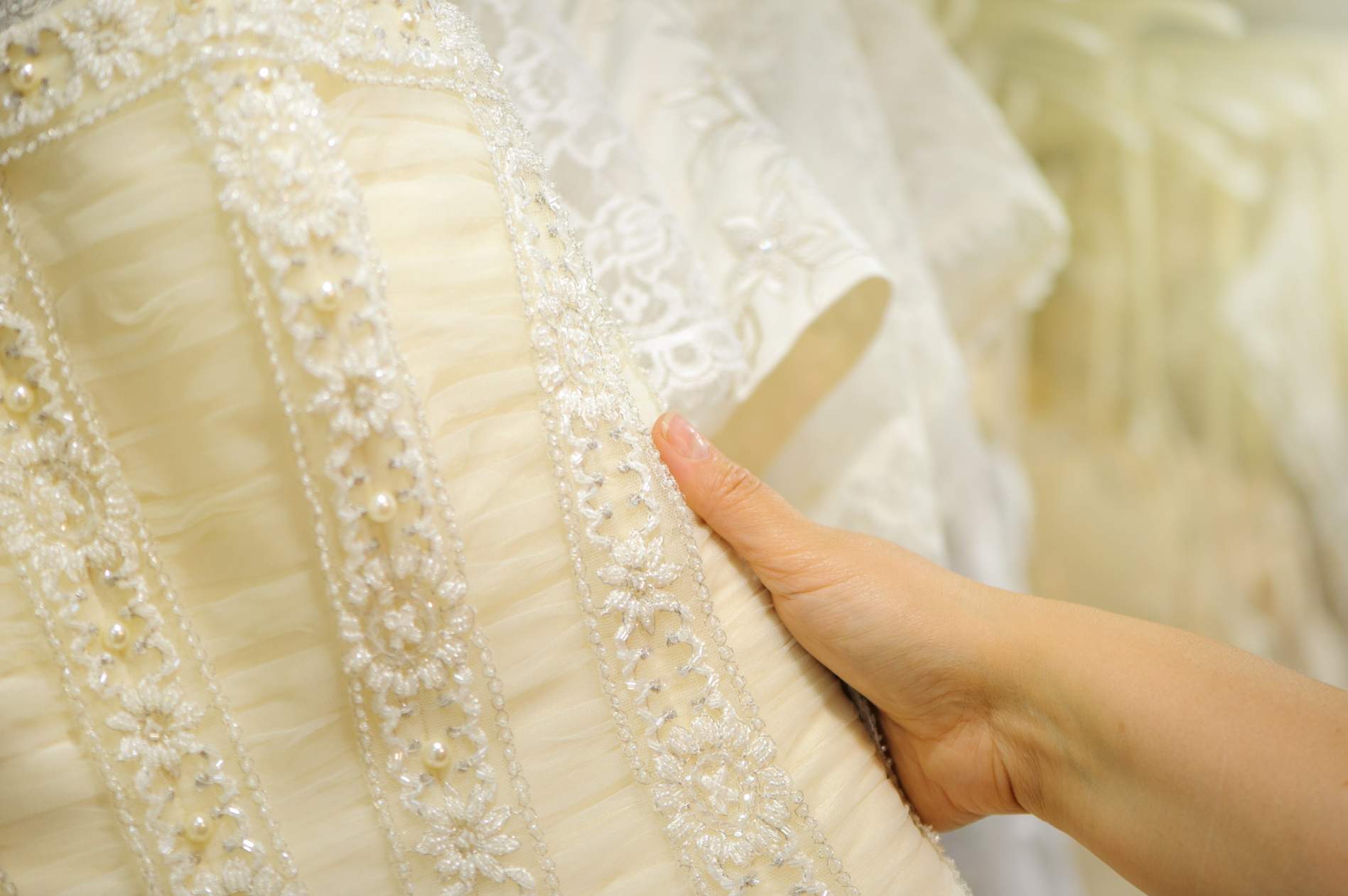 A close-up of a hand holding a white lace wedding dress adorned with intricate embroidery and pearl details. Other wedding dresses are visible in the background.